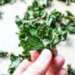 white hand holding a crispy kale chip with a background of kale chips on parchment paper