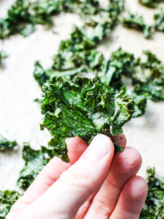 white hand holding a crispy kale chip with a background of kale chips on parchment paper