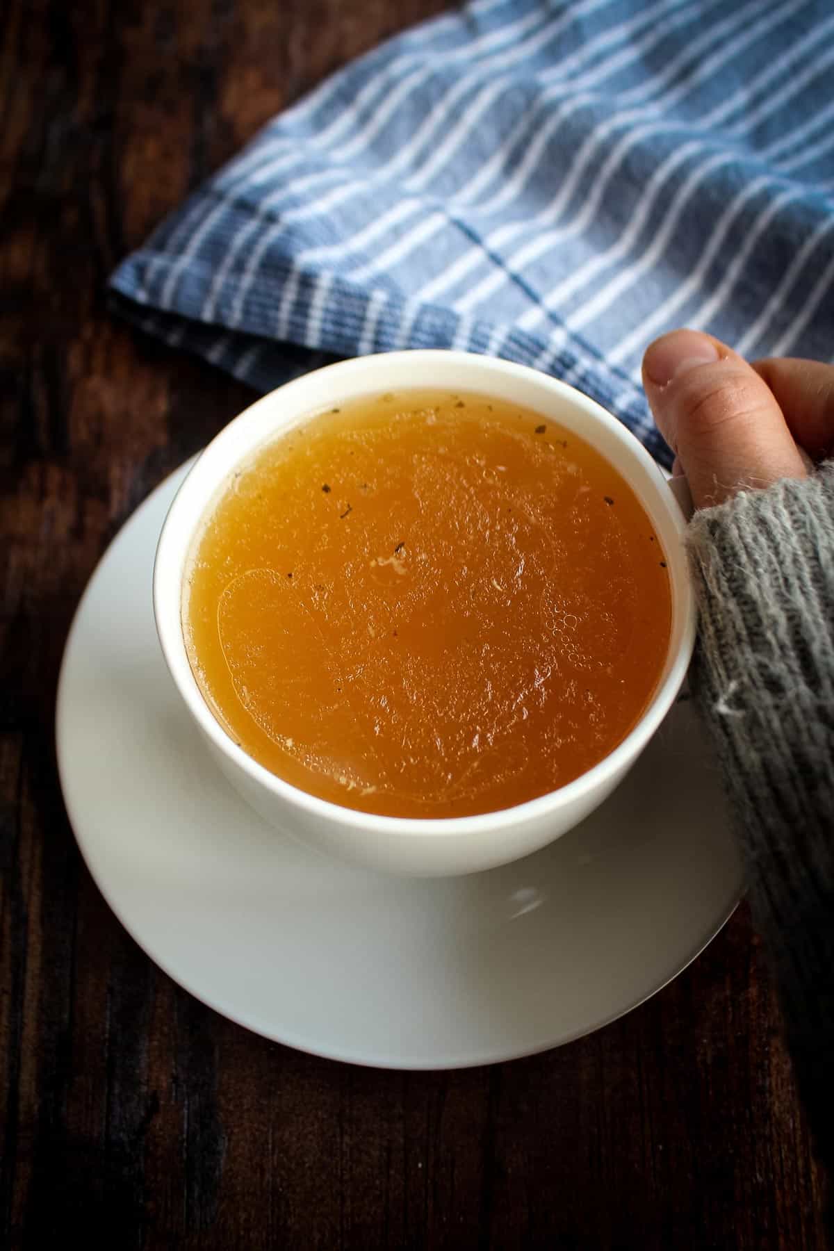 A cup of slow cooker chicken bone broth in a cup with a hand reaching to grab it.