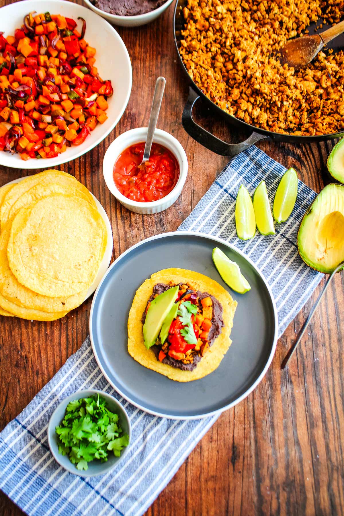 A single tempeh taco on a plate with toppings, tortillas, and a skillet of tempeh in the corner.