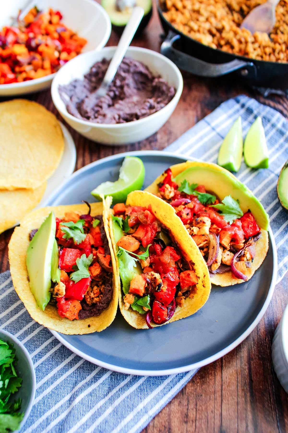 Three tempeh tacos on a blue plate with a bowl of black beans in the background.