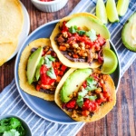 Three tempeh tacos on a plate with avocado and lime wedges.