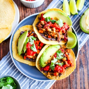 Three tempeh tacos on a plate with avocado and lime wedges.