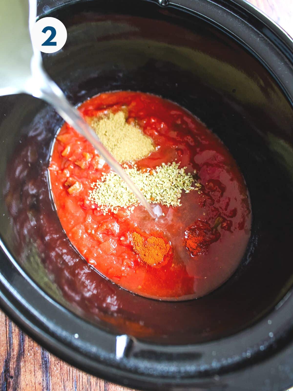 Tomato and seasonings in a crockpot with a pitcher adding some water.