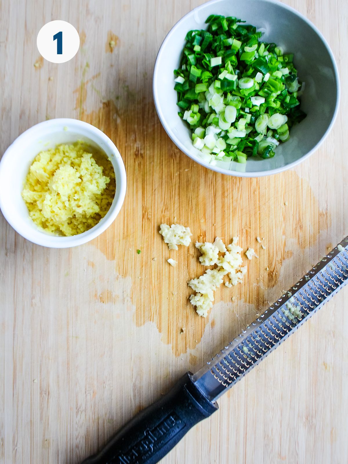 Grated ginger and minced green onions in a bowl with chopped garlic on a cutting board.