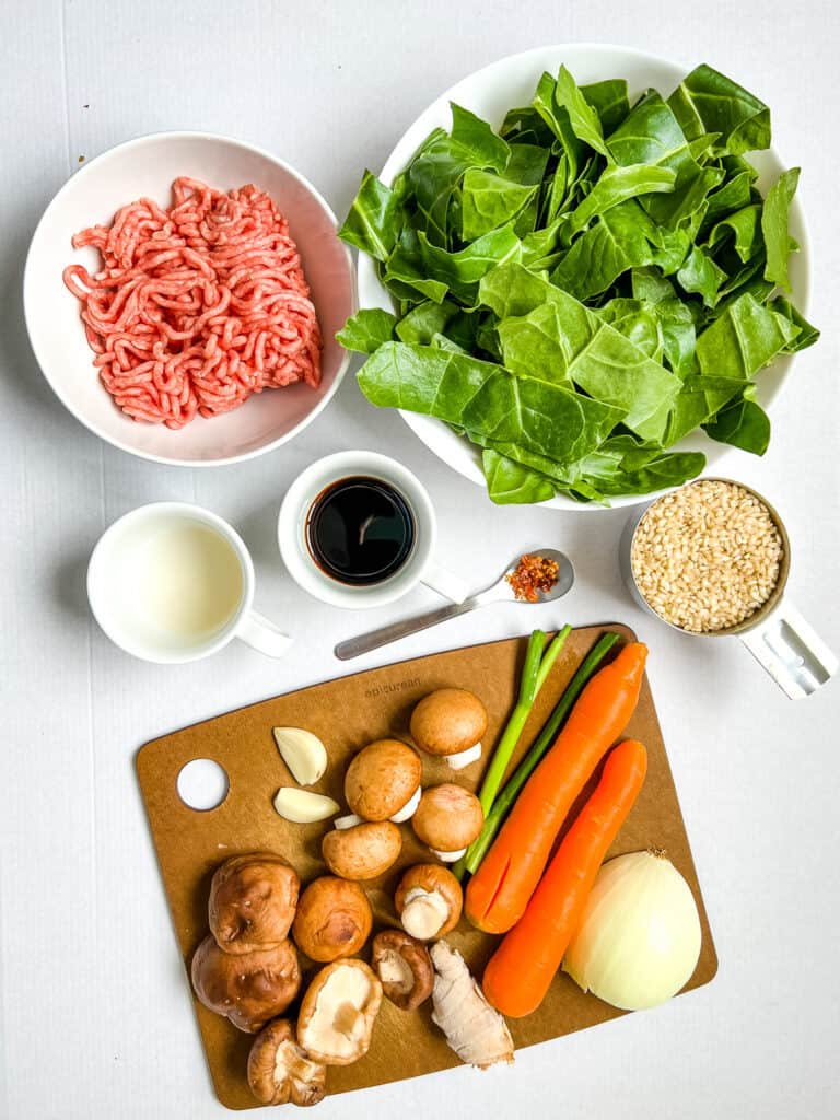 Ingredients displayed on white background - ground pork, chopped greens, uncooked brown rice, cutting board with half an onion, two carrots, a green onion, some crimini mushrooms and shiitake mushrooms, two cloves of garlic, a knob of ginger,, plus a small mug with vinegar and a small mug with soy sauce, and a small spoon with red pepper flakes. 