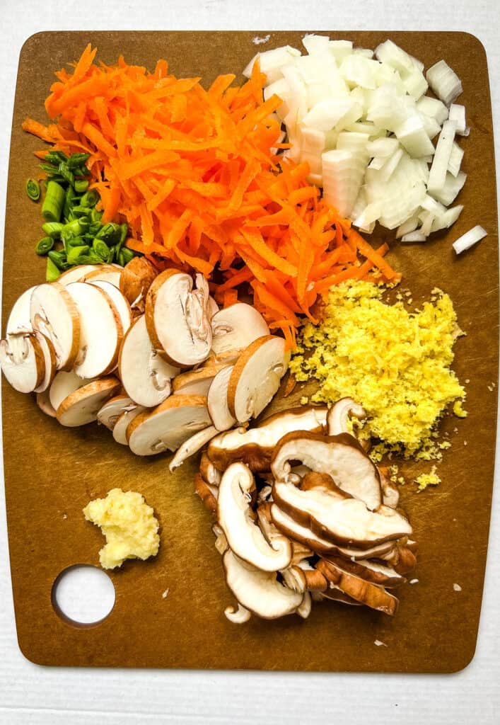 Cutting board with prepped vegetables, shredded carrots, diced onion, minced ginger, sliced mushrooms, crushed garlic, and sliced green onion.