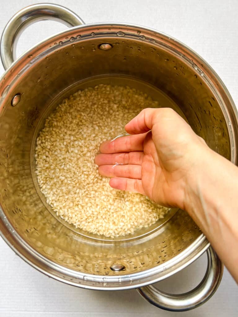 Measuring rice with a pot of brown rice, water, and hand showing the water coming up to the first knuckle of the middle finger, when placed mostly flat into the pot.