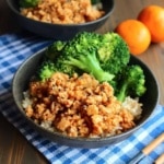 A bowl of ground orange chicken over rice and broccoli on the table with mandarins in the background along with another bowl.