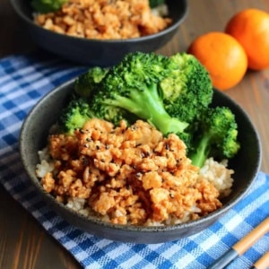 A bowl of ground orange chicken over rice and broccoli on the table with mandarins in the background along with another bowl.