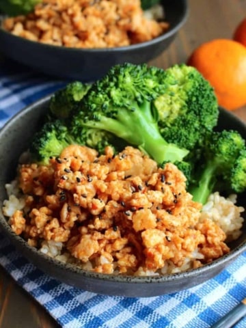 A bowl of ground orange chicken over rice and broccoli on the table with mandarins in the background along with another bowl.