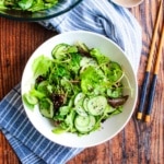 Sesame cucumber salad in a white bowl on the table with chopsticks to the right and a napkin underneath.