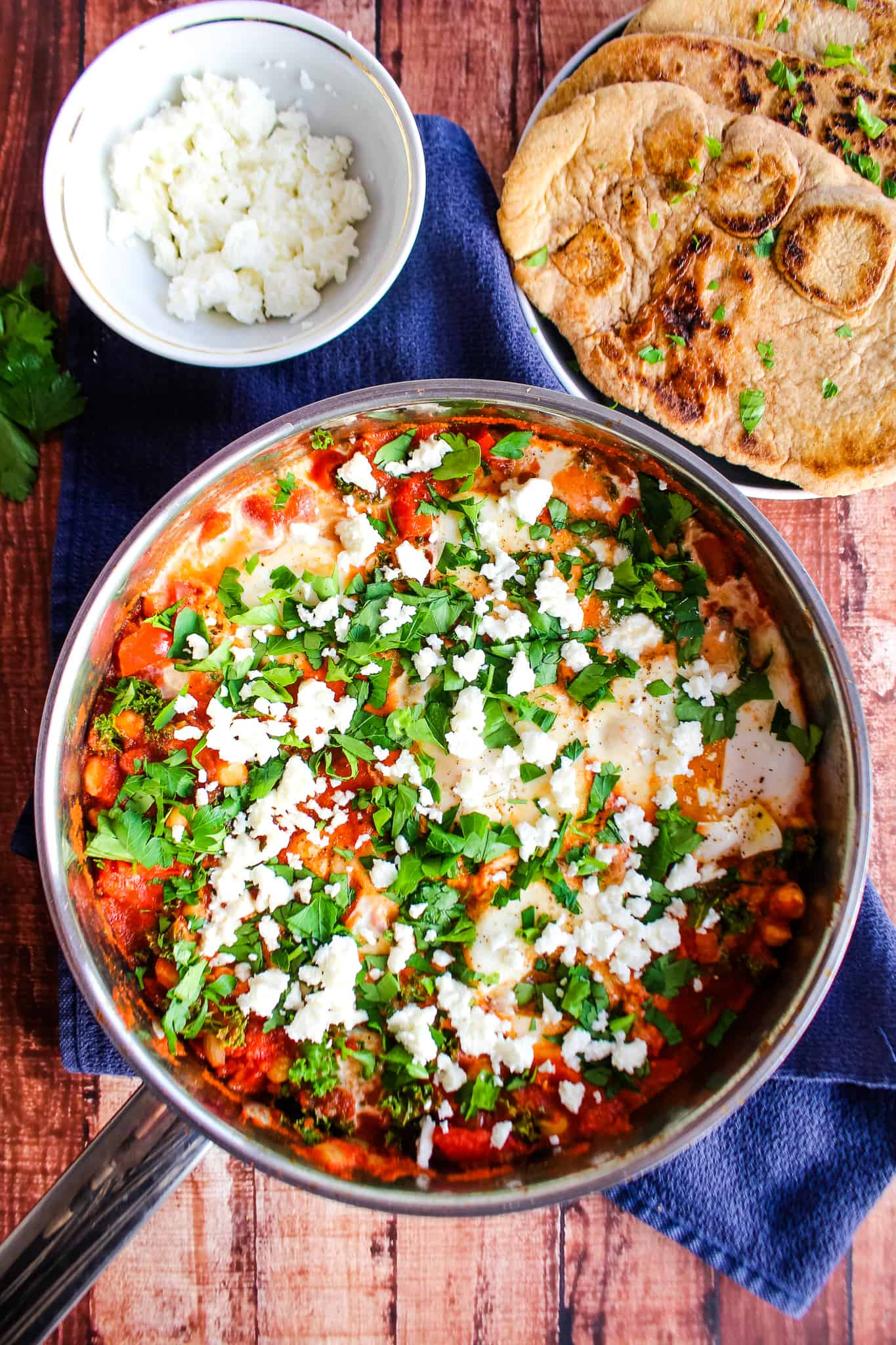 Shakshuka in a skillet topped with parsley and feta, next to a plate with buckwheat naan and a bowl of crumbled feta. 