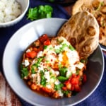 A gray bowl with a single serving shakshuka showing a bit of a runny egg, topped with feta and parsley, with a piece of buckwheat naan.