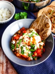 A gray bowl with a single serving shakshuka showing a bit of a runny egg, topped with feta and parsley, with a piece of buckwheat naan.