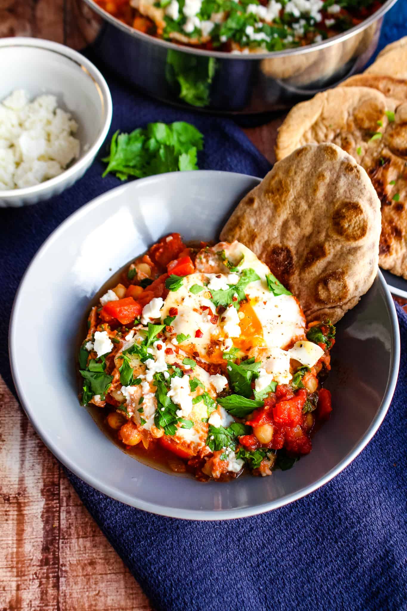 A gray bowl with a single serving shakshuka showing a bit of a runny egg, topped with feta and parsley, with a piece of buckwheat naan.