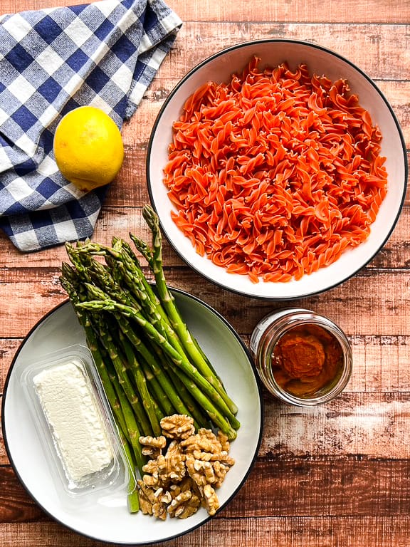 bowl of uncooked red lentil rotini, jar of sun dried tomatoes, uncooked whole asparagus spears, walnut halves, a log of goat cheese, and a lemon.