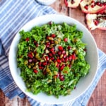 bowl of kale with pomegranate seeds and pumpkin seeds in foreground on blue napkin with opened pomegranate and bowl of pumpkin seeds in background.