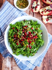 bowl of kale with pomegranate seeds and pumpkin seeds in foreground on blue napkin with opened pomegranate and bowl of pumpkin seeds in background.