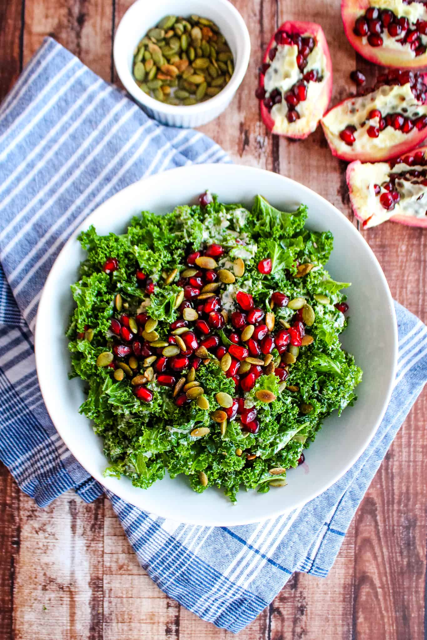 bowl of kale with pomegranate seeds and pumpkin seeds in foreground on blue napkin with opened pomegranate and bowl of pumpkin seeds in background
