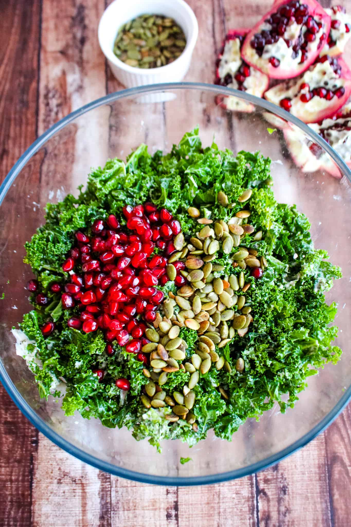 bowl of kale with pomegranate seeds and pumpkin seeds in foreground with opened pomegranate and bowl of pumpkin seeds in background.