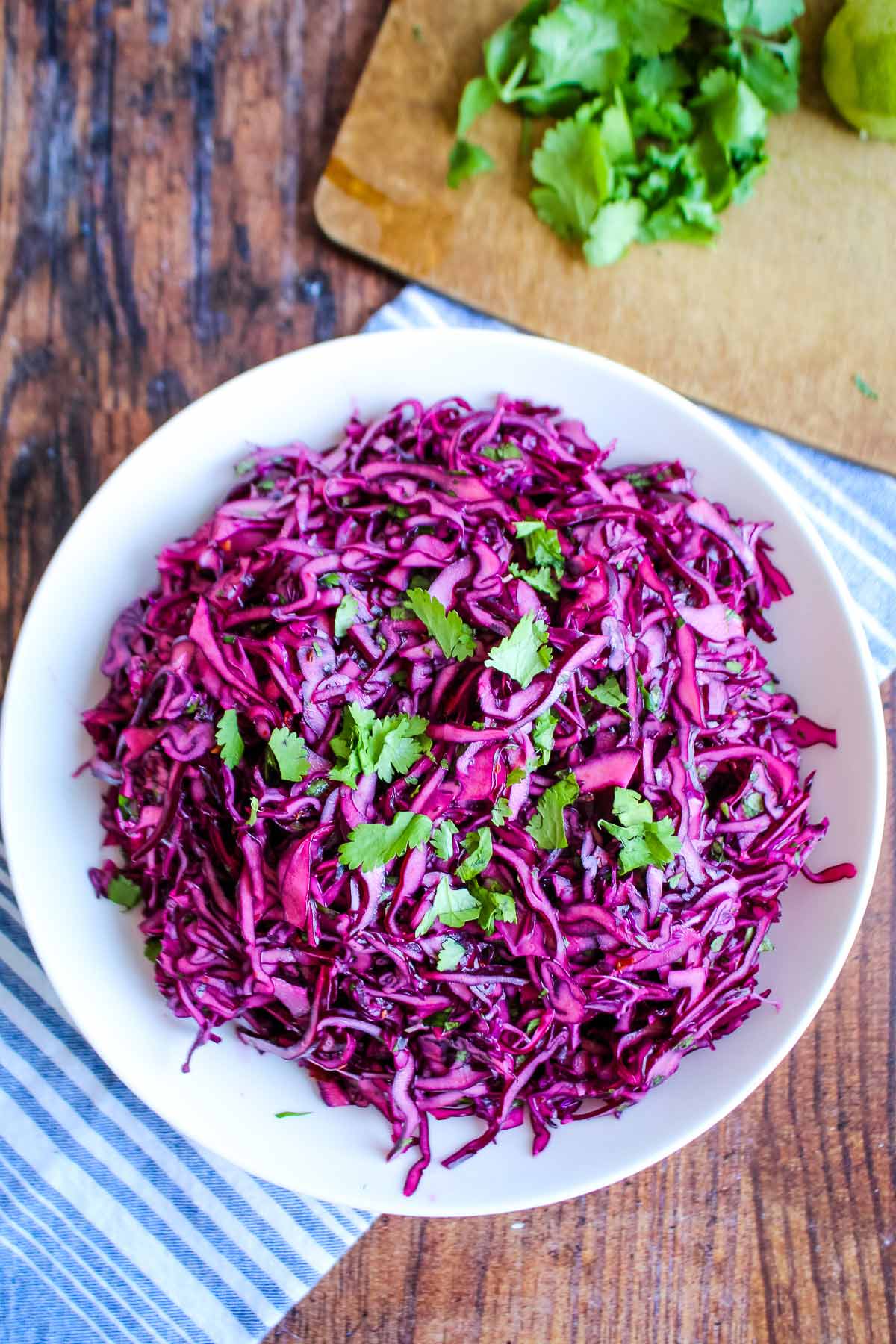 Red cabbage slaw in a white bowl on the table with chopped cilantro on a cutting board.