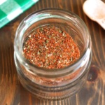 A jar of Italian sausage seasoning on the table with a small wooden spoon.