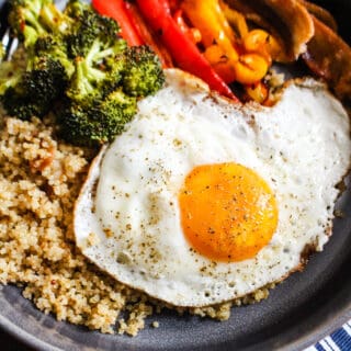 Close up bowl of quinoa, roasted broccoli, roasted bell peppers, and a fried egg.