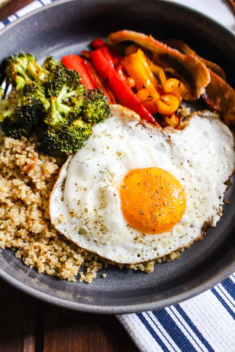 Close up bowl of quinoa, roasted broccoli, roasted bell peppers, and a fried egg.