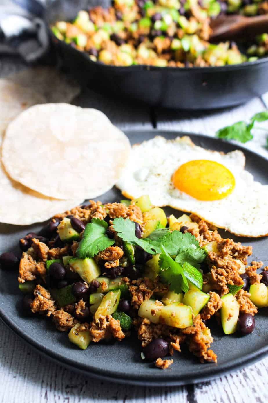 Slate colored plate with a hash of chorizo, zucchini, and black beans, with a side of mini tortillas and a sunny side up egg