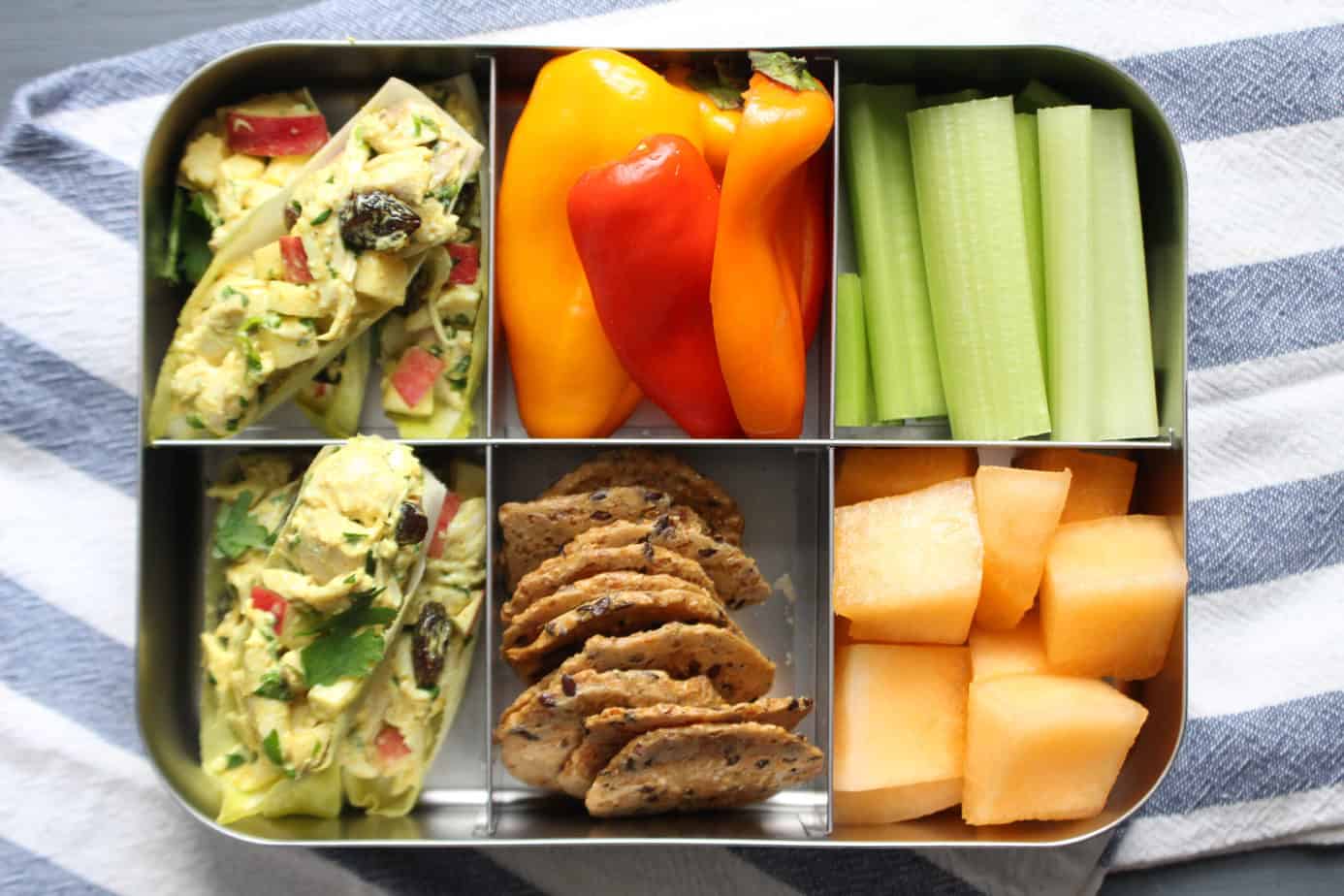 6-compartment metal lunch box with curry chicken salad endive boats, crackers, bell peppers, cantaloupe, and celery sticks