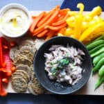 blue background with cutting board with hummus, crackers, canned mackerel, and colorful vegetables
