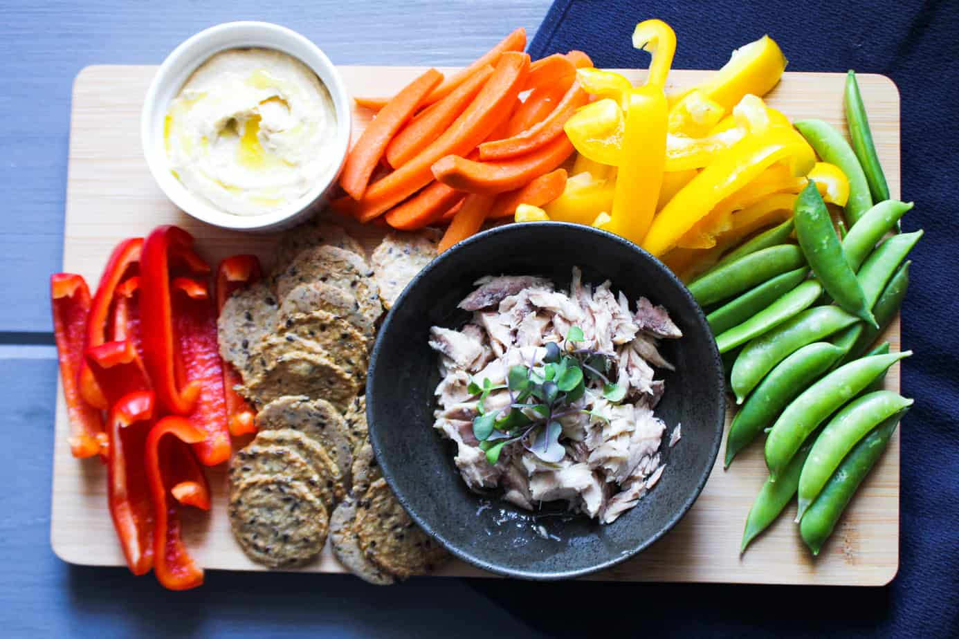 blue background with cutting board with hummus, crackers, canned mackerel, and colorful vegetables