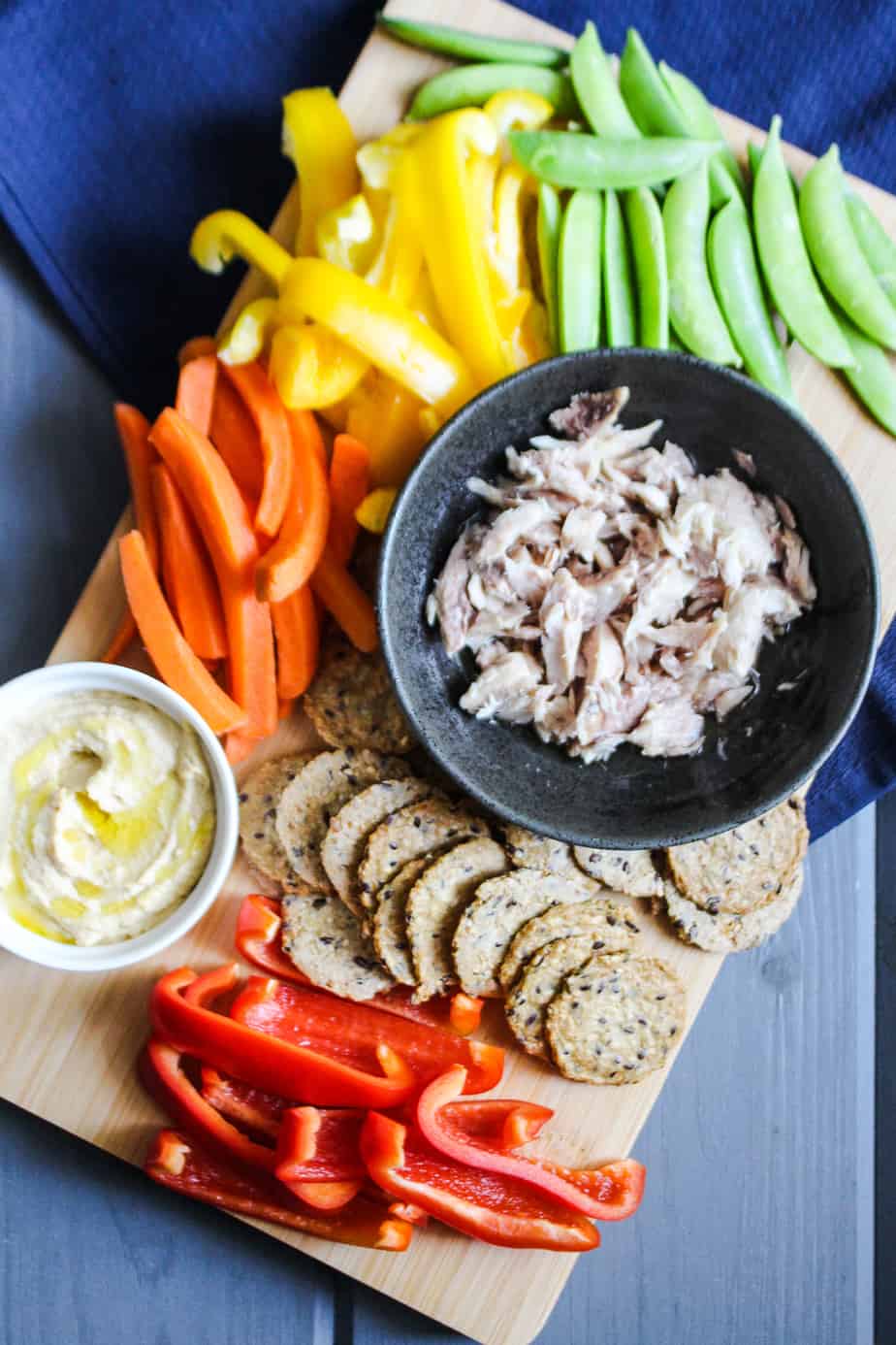 blue background with cutting board with hummus, crackers, canned mackerel, and colorful vegetables