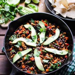 skillet with colorful vegetables, beans, turkey, and decorative slices of avocado with chips, salsa, and cilantro