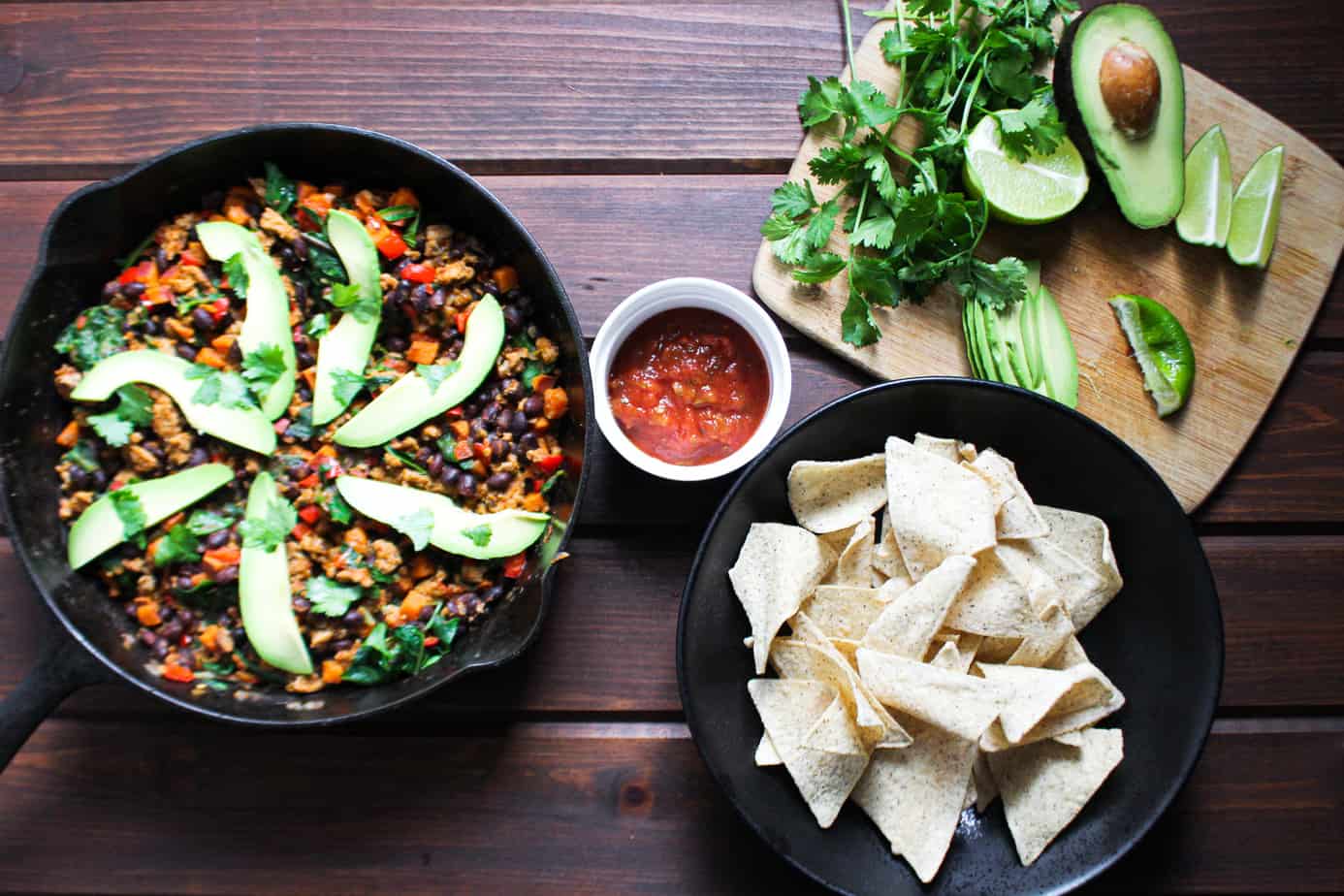 skillet with colorful vegetables, beans, turkey, and decorative slices of avocado with chips, salsa, and cilantro