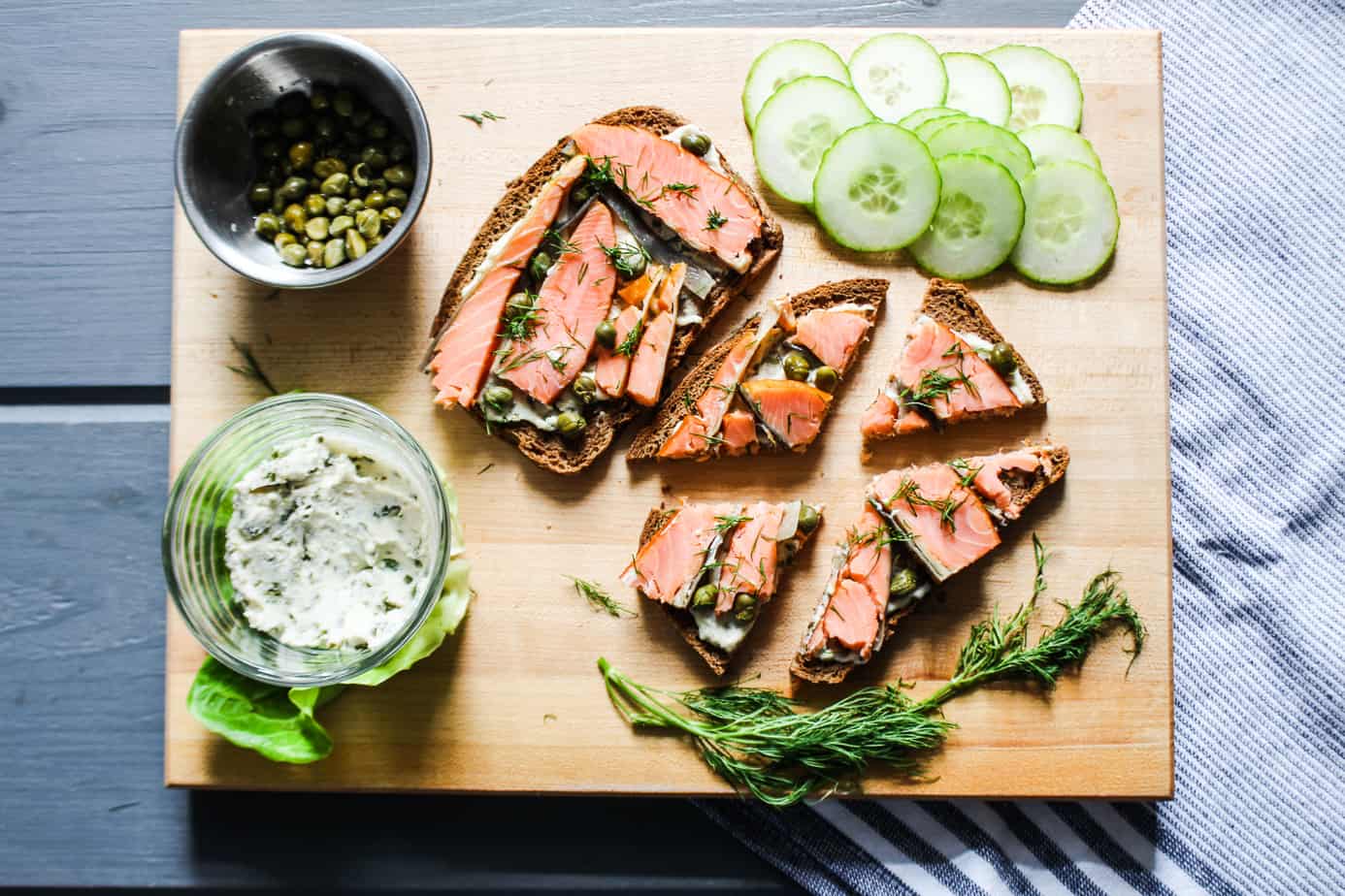 cutting board with smoked salmon toasts, sliced cucumbers, a sprig of fresh dil, and containers of capers and chive cream cheese