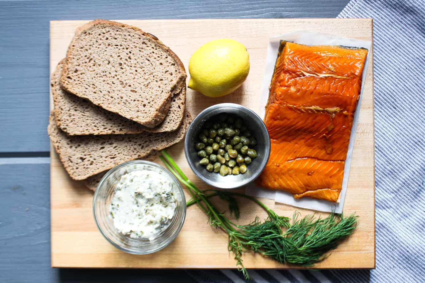 cutting board with 4 slices of dark bread, a fillet of smoked salmon, capers, a lemon, fresh dill, and cream cheese studded with chives