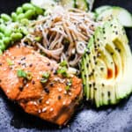 close up of black bowl with salmon, soba, edamame, cucumbers, avocado, black and white sesame seeds