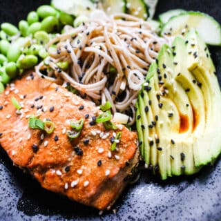 close up of black bowl with salmon, soba, edamame, cucumbers, avocado, black and white sesame seeds