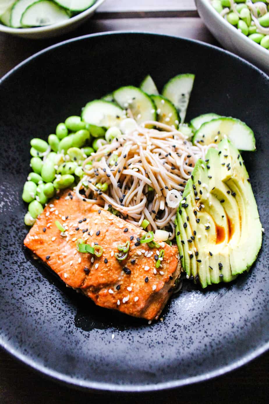 close up of black bowl with salmon, soba, edamame, cucumbers, avocado, black and white sesame seeds