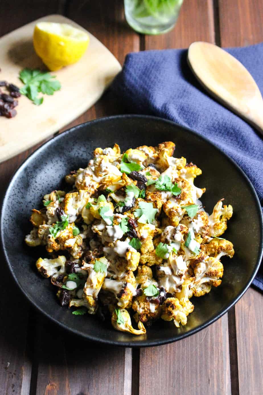 black bowl filled with yellow tinted curry roasted cauliflower combined with raisins, cilantro, and lemon tahini sauce
