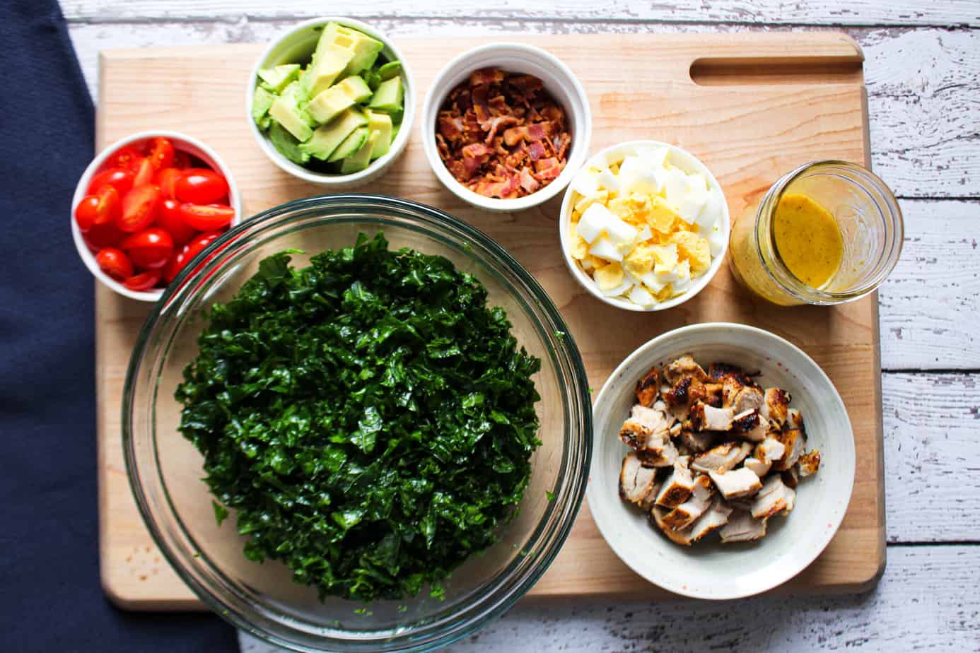 chopped ingredients on cutting board: avocado, bacon, grape tomatoes, kale, cooked chicken, eggs, dressing