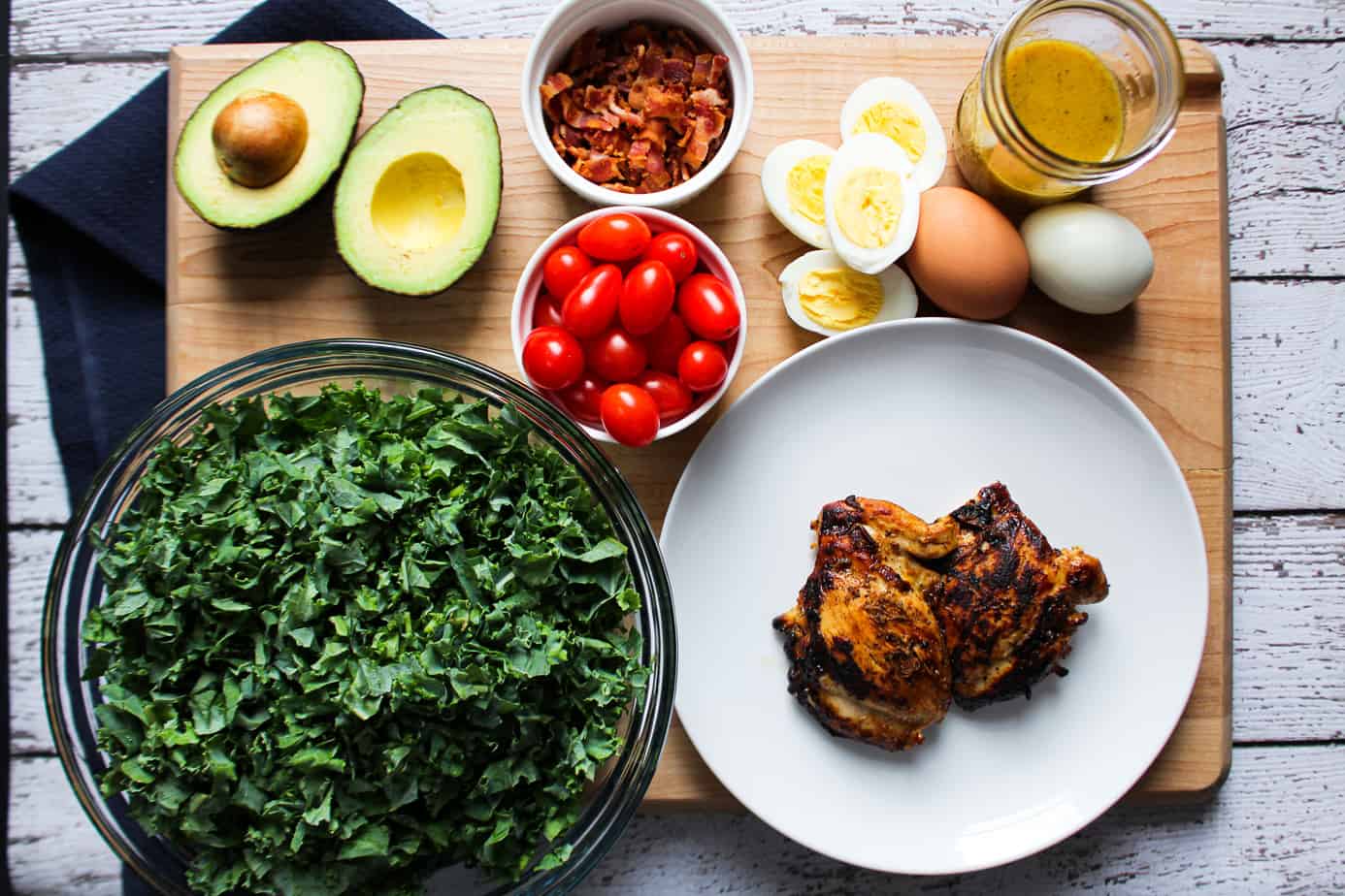 ingredients on cutting board: avocado, bacon, grape tomatoes, kale, cooked chicken, eggs, dressing