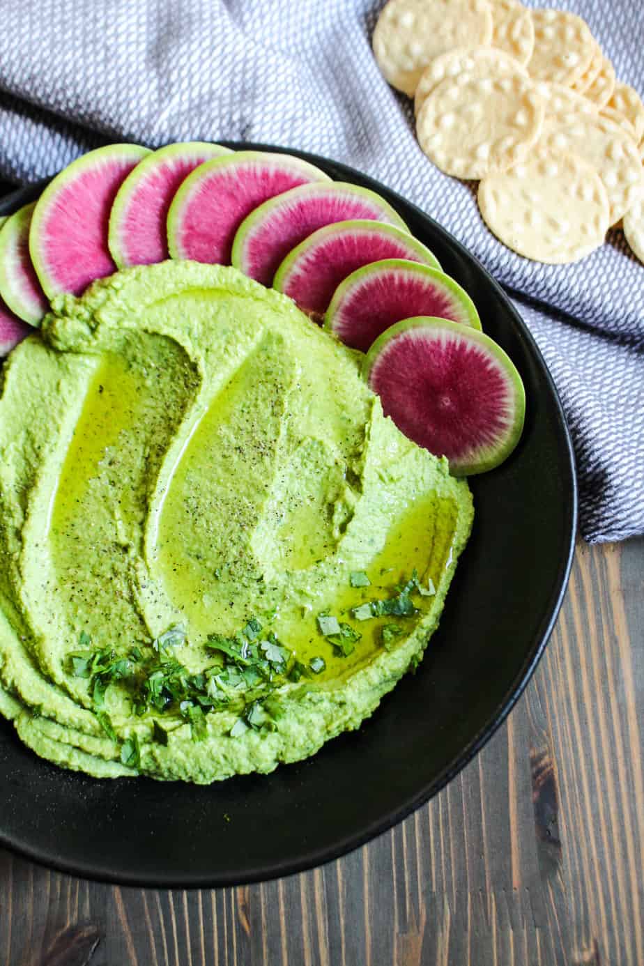 black bowl with green hummus, garnished with olive oil and herbs, watermelon radish slices, and some rice crackers on the side