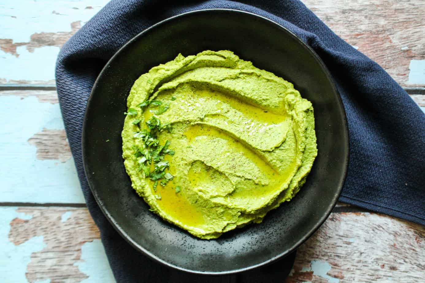 black bowl with green hummus, garnished with olive oil and herbs