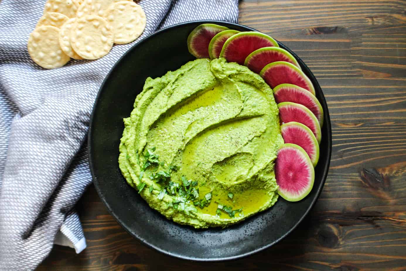 black bowl with green hummus, garnished with olive oil and herbs, watermelon radish slices, and some rice crackers on the side