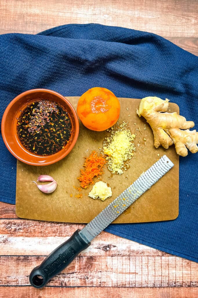 Cutting board with sauce ingredients, piece of ginger, garlic, and orange, with grated ginger, garlic, and orange zest in the center next to microplane grater.