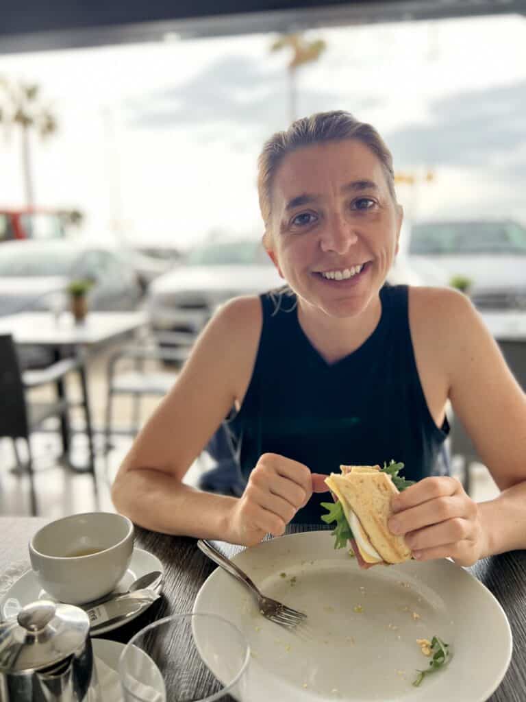 a woman in a black tank top, post-run, smiling and holding half a GF pitufo sandwich with arugula, tomato, egg, jamon, and avocado