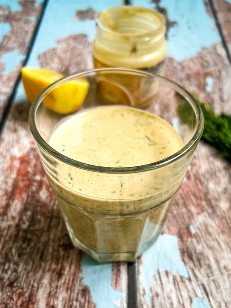 glass with mixed tahini dressing in the foreground, half a lemon, jar of tahini, and fresh dill in the background